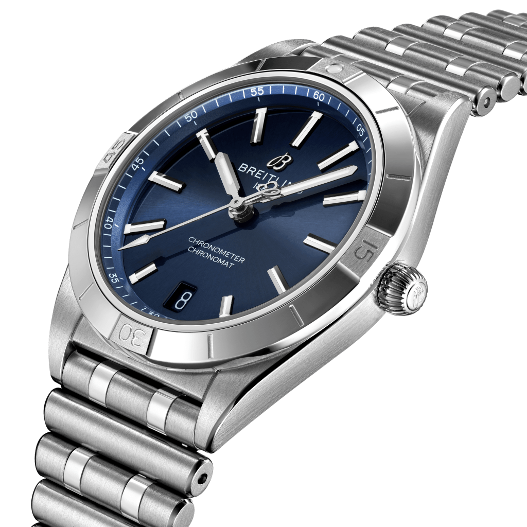 Relógio de pulso em aço inoxidável com um mostrador azul, marcadores de hora e ponteiros prateados, uma janela de data às 3 horas e o nome da marca ‘Breitling’ abaixo da posição das 12 horas. O relógio tem uma pulseira de elos e uma coroa canelada no lado direito. O horário no relógio lê aproximadamente 10:10.