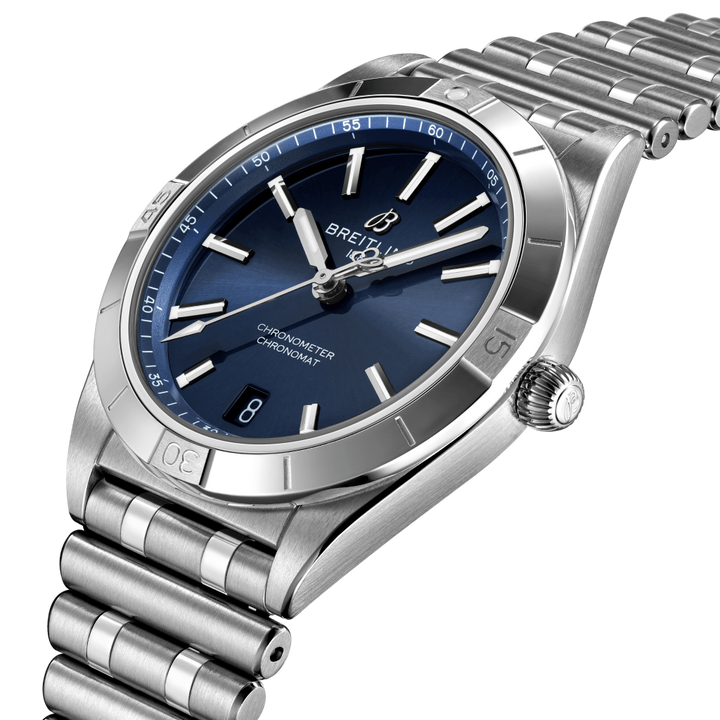 Relógio de pulso em aço inoxidável com um mostrador azul, marcadores de hora e ponteiros prateados, uma janela de data às 3 horas e o nome da marca ‘Breitling’ abaixo da posição das 12 horas. O relógio tem uma pulseira de elos e uma coroa canelada no lado direito. O horário no relógio lê aproximadamente 10:10.