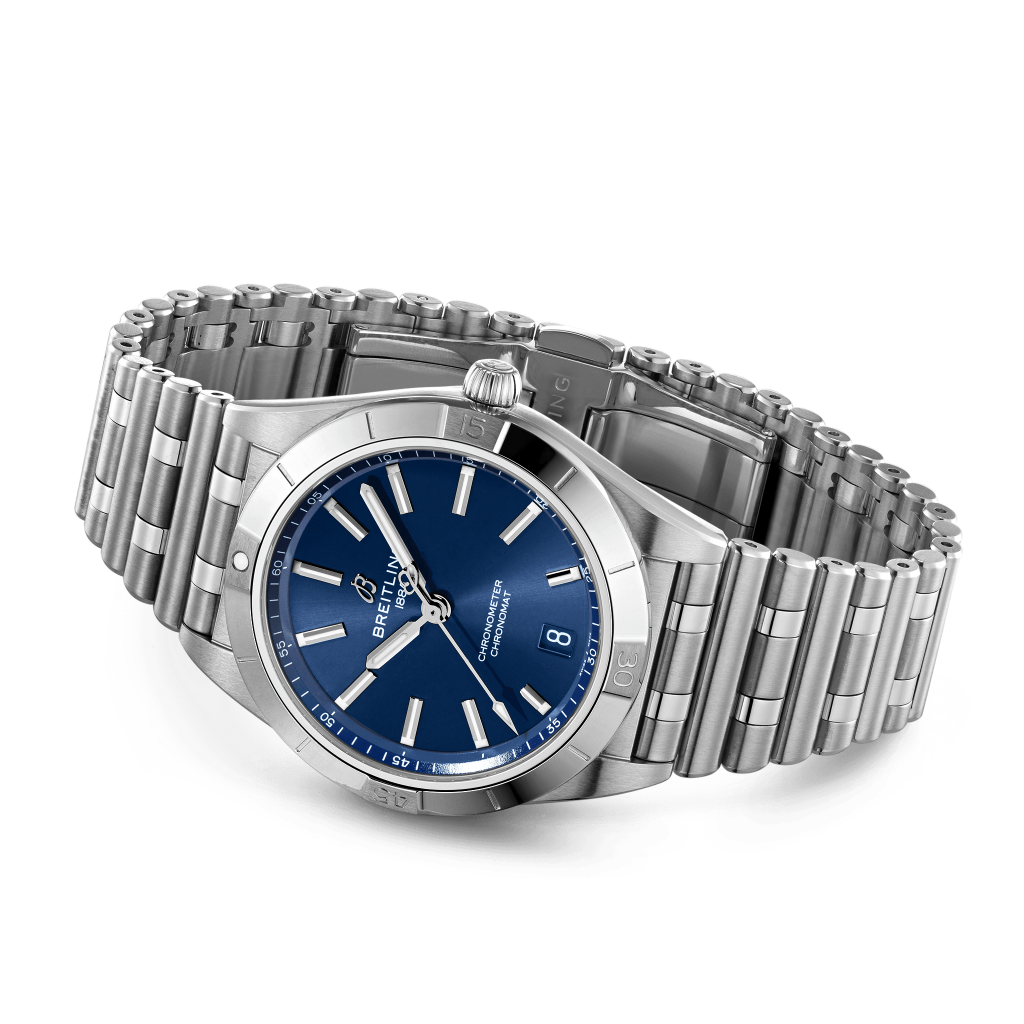 Um relógio de pulso em aço inoxidável com uma pulseira de elos. O relógio tem um mostrador azul com ponteiros e marcadores de hora em tom prateado. Há uma exibição de data na posição das 3 horas, e o nome da marca é visível abaixo do marcador das 12 horas. A coroa do relógio está posicionada no lado direito da caixa, e parece ter um acabamento polido.