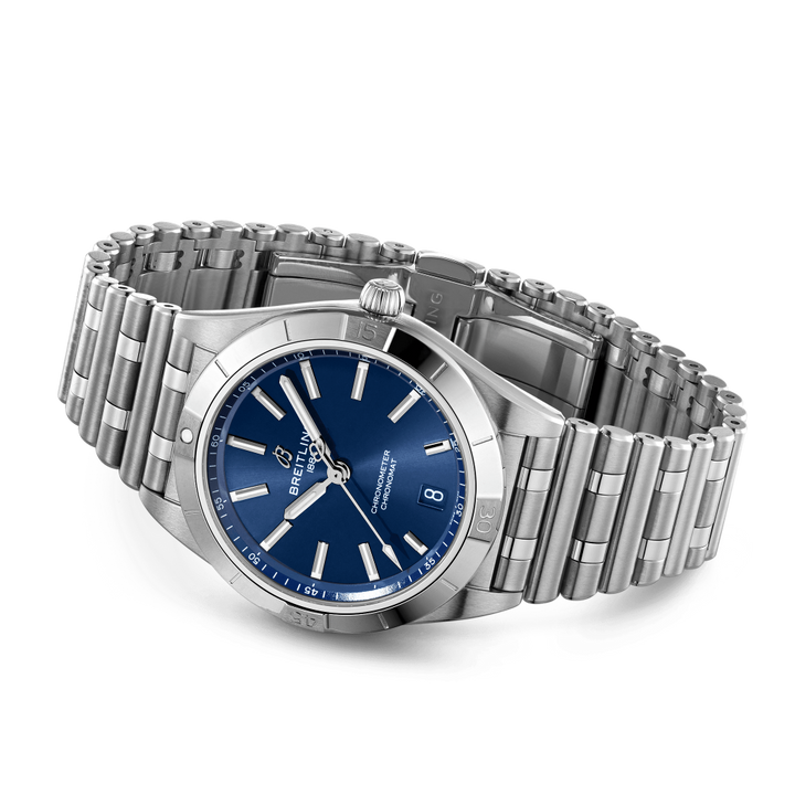 Um relógio de pulso em aço inoxidável com uma pulseira de elos. O relógio tem um mostrador azul com ponteiros e marcadores de hora em tom prateado. Há uma exibição de data na posição das 3 horas, e o nome da marca é visível abaixo do marcador das 12 horas. A coroa do relógio está posicionada no lado direito da caixa, e parece ter um acabamento polido.