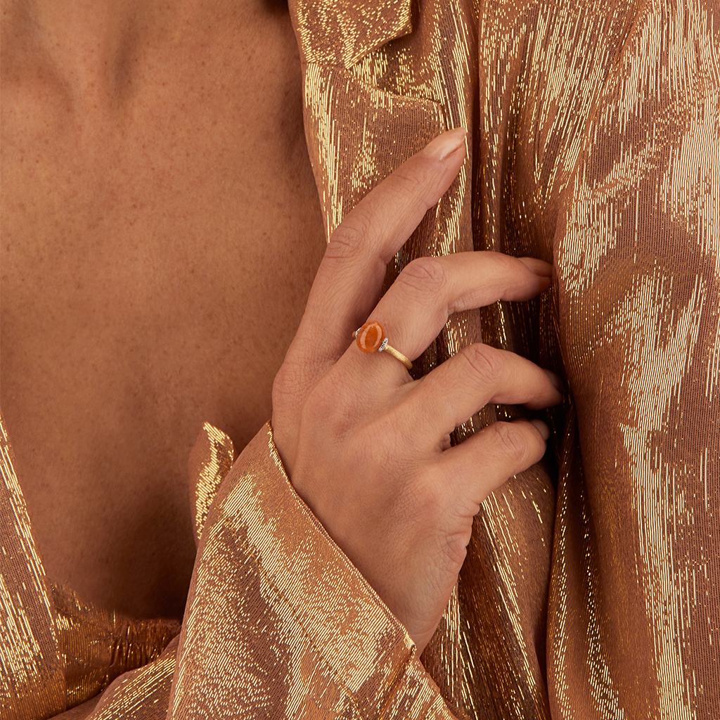 imagem de mão de senhora a segurar camisola e a usar no dedo do meio o Anel amuleti Petra com uma aventurina laranja em aro de ouro 18k e rodeada de diamantes