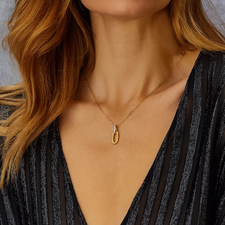 Uma pessoa usando um colar delicado de corrente de ouro com um pequeno pingente brilhante incrustado de diamantes que se assemelha a um laço ou nó estilizado. O colar é exibido contra o pano de fundo de uma blusa de cor escura com um decote V profundo, que complementa a elegância da joia. O cabelo da pessoa é castanho-avermelhado e cai suavemente sobre os ombros, enquadrando o colar e chamando a atenção para ele.