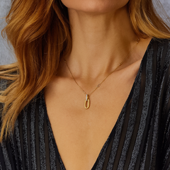 Uma pessoa usando um colar delicado de corrente de ouro com um pequeno pingente brilhante incrustado de diamantes que se assemelha a um laço ou nó estilizado. O colar é exibido contra o pano de fundo de uma blusa de cor escura com um decote V profundo, que complementa a elegância da joia. O cabelo da pessoa é castanho-avermelhado e cai suavemente sobre os ombros, enquadrando o colar e chamando a atenção para ele.