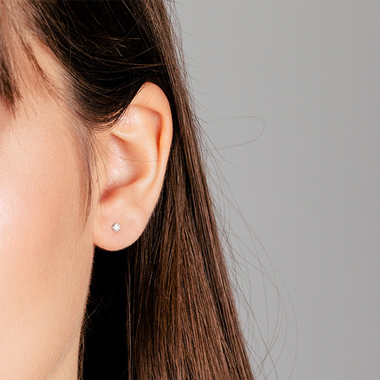 Close-up da orelha de uma mulher de cabelo castanho escuro usando um pequeno brinco de pino brilhante. O fundo é de tom cinza neutro, contrastando com a pele quente e o cabelo escuro da pessoa.