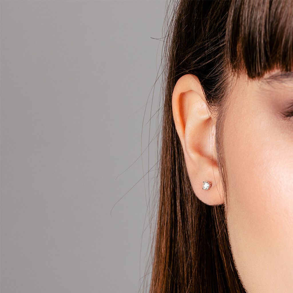 Close-up de uma orelha usando um brinco de pino brilhante, com parte do cabelo visível. O fundo é cinza e simples, destacando o brinco elegante.