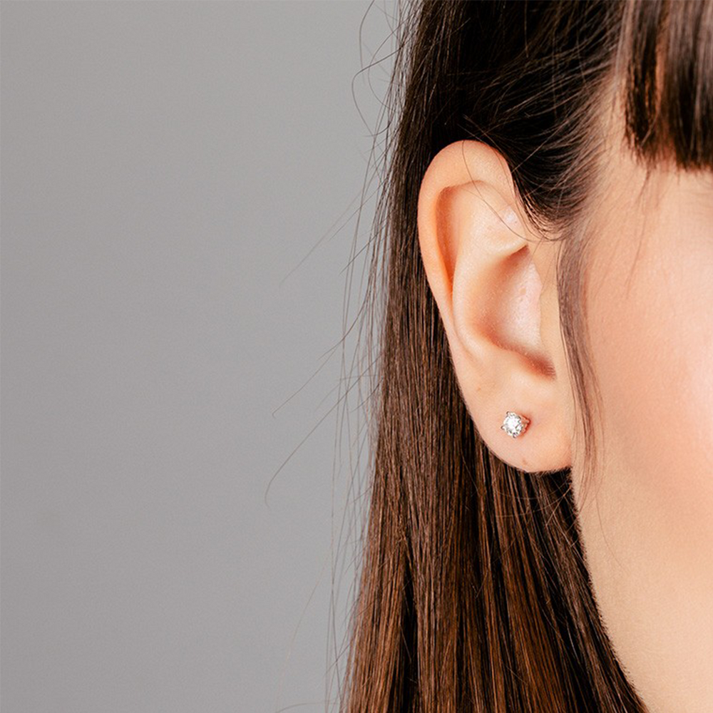 Close-up de uma orelha de uma mulher usando um brinco luxuoso feito de ouro com um proeminente diamante central. O design do brinco é simples, mas elegante, apresentando o diamante em uma configuração minimalista de ouro que destaca seu brilho contra o fundo cinza neutro. O cabelo castanho reto da mulher está preso atrás da orelha, atraindo a atenção para a joia cintilante.