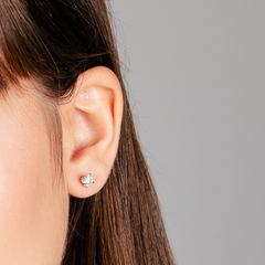 Close-up da orelha de uma mulher usando um brinco de diamante brilhante contra um fundo cinza. O brinco está posicionado no lóbulo da orelha, captando a luz e mostrando as facetas da pedra preciosa. A imagem foca no brinco, proporcionando uma visão clara do seu design e de como fica na orelha