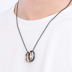 Colar de aço da Emporio Armani com um pingente de dois anéis entrelaçados, um com acabamento brilhante e o outro fosco. A corrente é de aço escurecido com elos pequenos e uniformes. O colar é exibido sobre uma camiseta branca, destacando o design elegante da joia.