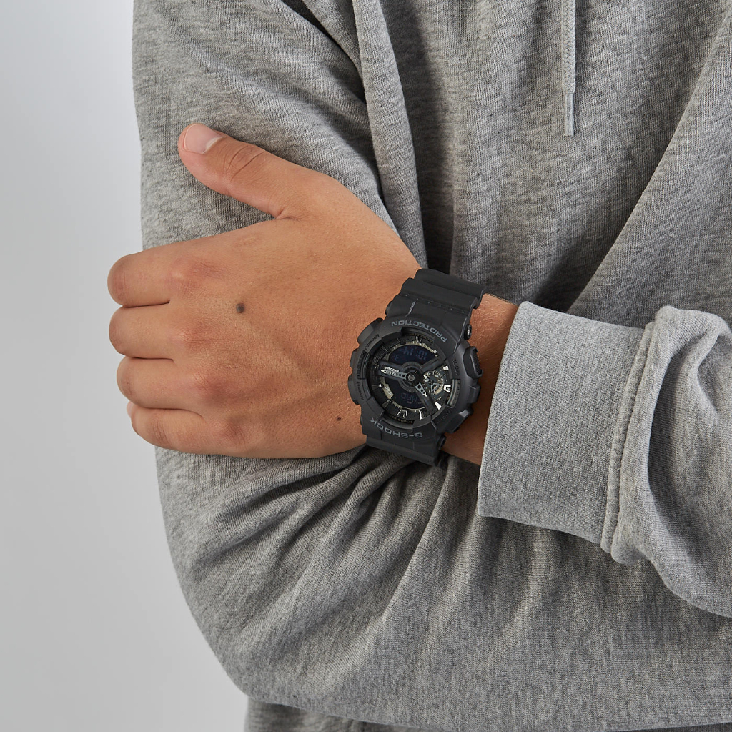 Um relógio de pulso de resina preta no pulso esquerdo de uma pessoa vestindo uma camisa de manga comprida cinza. O relógio possui vários mostradores e botões, indicando que pode ter várias funções além de mostrar as horas. O braço da pessoa está dobrado no cotovelo, e o fundo é de uma cor neutra clara, destacando os detalhes escuros do relógio.