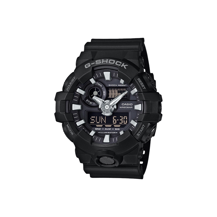 Relógio Casio G-Shock com bracelete de resina preta. O mostrador combina displays analógicos e digitais, com ponteiros prateados e marcadores de hora sobre um fundo preto. O mostrador digital exibe a hora em formato numérico. O bisel é marcado com indicadores de minutos e a inscrição “SHOCK RESIST”. Funções adicionais incluem exibição de dia e data, além de configurações de alarme visíveis no mostrador.