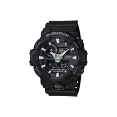 Relógio Casio G-Shock com bracelete de resina preta. O mostrador combina displays analógicos e digitais, com ponteiros prateados e marcadores de hora sobre um fundo preto. O mostrador digital exibe a hora em formato numérico. O bisel é marcado com indicadores de minutos e a inscrição “SHOCK RESIST”. Funções adicionais incluem exibição de dia e data, além de configurações de alarme visíveis no mostrador.
