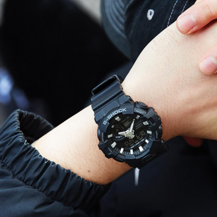 Relógio Casio G-Shock de resina preta no pulso esquerdo de uma pessoa. O relógio tem um design robusto com múltiplos botões e uma pulseira texturizada. O mostrador é detalhado, apresentando ponteiros analógicos e displays digitais, indicando sua funcionalidade dupla. O fundo está desfocado, destacando o relógio.