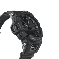 relógio Casio G-Shock de resina preta. O design é robusto, com múltiplos botões e detalhes texturizados nas laterais. O display é digital, com indicadores para vários modos como alarme, cronômetro e hora mundial. A palavra ‘PROTECTION’ está destacada acima da tela digital, enfatizando a durabilidade do relógio. A pulseira parece ser feita de um material durável, provavelmente borracha ou um material sintético similar, adequado para uso ativo ou ao ar livre.