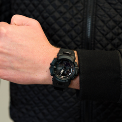 Pulso usando um relógio de resina preta. O relógio possui um display digital-analógico com múltiplos mostradores e botões laterais. O fundo está desfocado, destacando o relógio, que é o ponto focal da imagem. A pessoa está vestindo uma jaqueta escura com padrão acolchoado, sugerindo um ambiente casual e estiloso
