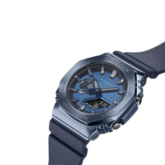 Relógio de pulso de resina com mostrador transparente azul e pulseira azul correspondente. O design é robusto, com parafusos visíveis no aro, e a marca ‘G-SHOCK’ está destacada na parte superior do mostrador. Os ponteiros são metálicos e há várias marcações e textos no mostrador indicando recursos de medição de tempo. O fundo é branco, destacando os detalhes do relógio.
