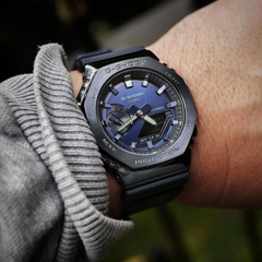 “Imagem de close-up de um pulso usando um relógio G-Shock de resina preta. O relógio tem um mostrador digital-analógico com detalhes em azul no mostrador e é rotulado com o logotipo G-Shock, nome do modelo e o texto ‘PROTECTION’. A pessoa está usando uma roupa de manga comprida cinza, e o fundo parece ser natural e desfocado, possivelmente indicando um ambiente ao ar livre.