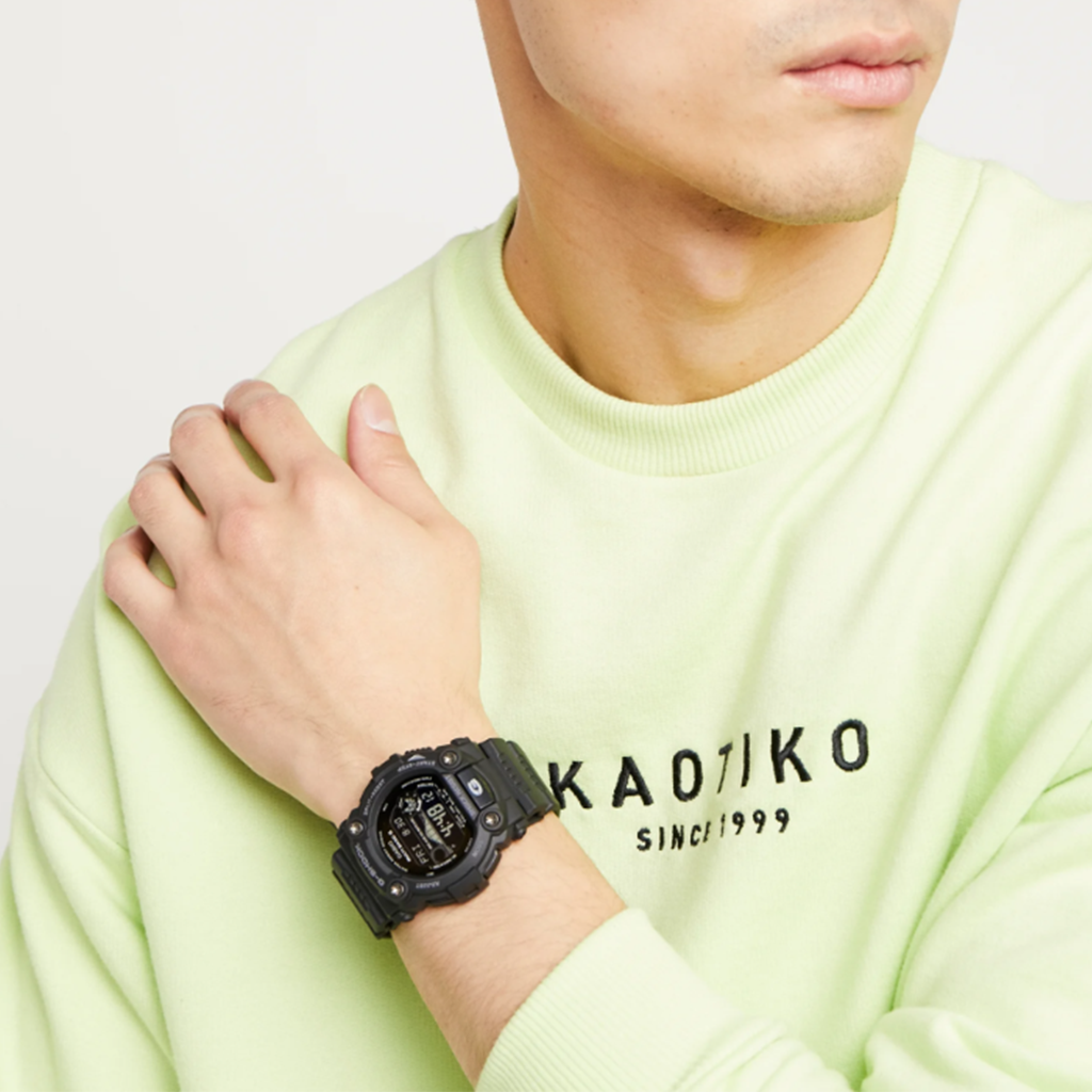 Um relógio de pulso de resina preta usado no pulso esquerdo de uma pessoa vestindo uma camisa de manga comprida verde-clara com ‘KAOTIKO SINCE 1999’ impresso no peito. O fundo é claro e simples, destacando o relógio e o texto da camisa