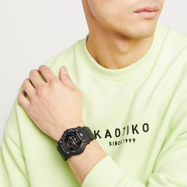 Um relógio de pulso de resina preta usado no pulso esquerdo de uma pessoa vestindo uma camisa de manga comprida verde-clara com ‘KAOTIKO SINCE 1999’ impresso no peito. O fundo é claro e simples, destacando o relógio e o texto da camisa