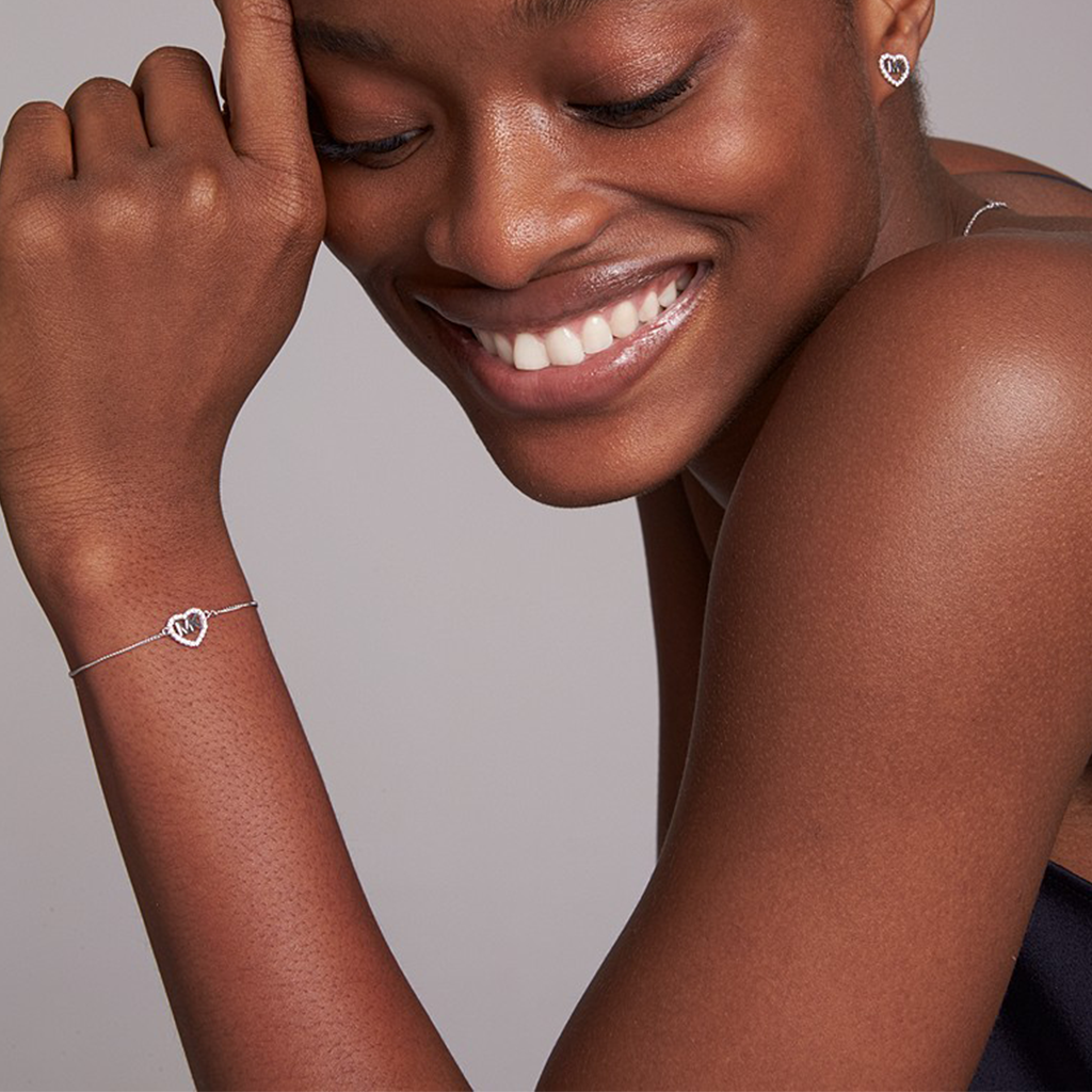A imagem apresenta um close-up do braço esquerdo de uma mulher levantado ao nível do ombro, com a mão descansando suavemente perto da área do pescoço. O foco está em uma pulseira delicada usada no pulso, que tem um design simples, mas elegante, apresentando uma forma de coração. O tom de pele da pessoa é escuro, e ela está usando o que parece ser um brinco de prata em forma de coração que combina com a pulseira. 