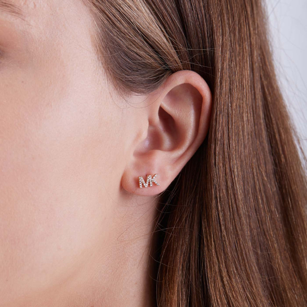 Close-up da orelha esquerda de uma pessoa usando um pequeno brinco brilhante em forma das iniciais ‘MK’. O fundo é de um tom cinza neutro, e o foco está no brinco, que se destaca devido ao seu brilho contra a pele da pessoa e o cabelo castanho claro preso atrás da orelha