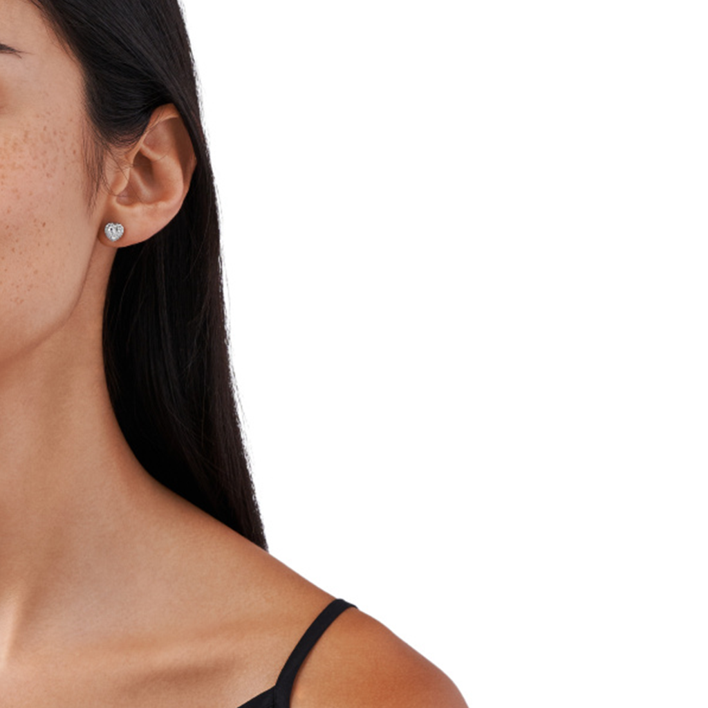 Imagem em close-up do lóbulo da orelha de uma mulher com um único brinco de diamante redondo. O brinco cintila de forma sutil e está fixado num estilo simples e clássico que complementa a forma da orelha. O cabelo da pessoa é escuro e liso, preso atrás da orelha para destacar o brinco de forma proeminente. Apenas parte do perfil da pessoa é visível, com foco no brinco como o principal assunto de interesse.