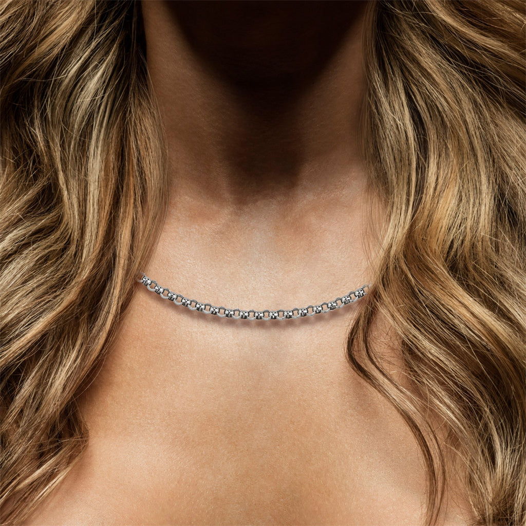 Imagem em close-up de um colar de prata Tashi usado ao redor do pescoço. O colar apresenta elos intricados e uniformemente espaçados que refletem a luz, conferindo um brilho delicado contra a pele. O fundo é uma pessoa com cabelo loiro e ondulado caindo sobre os ombros, proporcionando um contraste natural que destaca a elegância da joia.