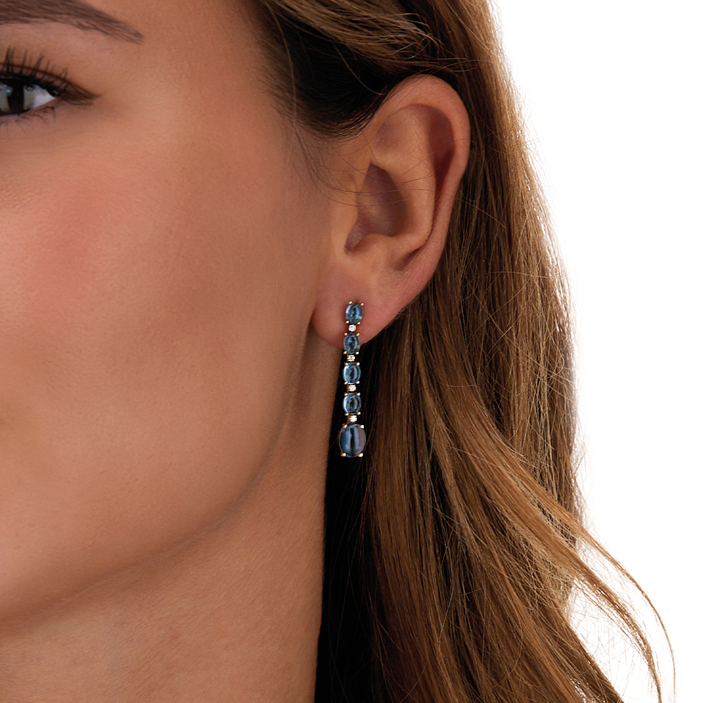 Uma visão em close-up da orelha direita de uma mulher exibindo um brinco longo. O brinco é adornado com topázios e pende elegantemente do lóbulo da orelha. O cabelo da mulher é castanho claro com ondas suaves caindo.