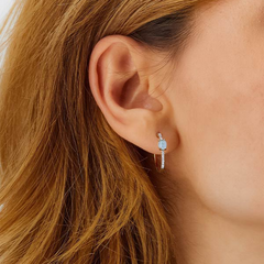 Close-up da orelha de uma mulher com cabelo castanho, usando um brinco que apresenta uma pedra preciosa azul no topo e um elemento pendente abaixo. O fundo é indistinto, focando a atenção no design do brinco e na forma como ele complementa a cor do cabelo do usuário.