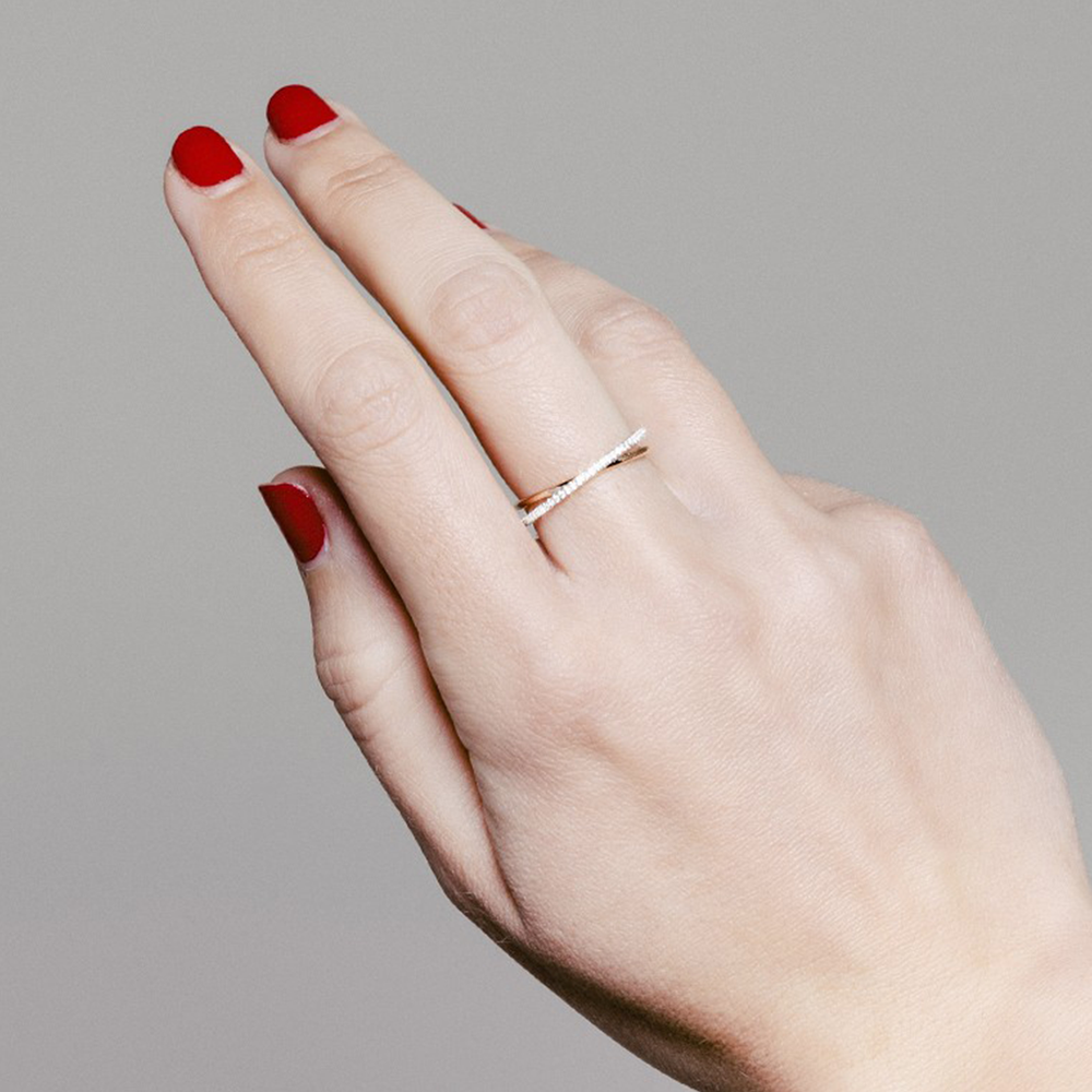 Uma imagem em close-up de uma mão com o dedo anelar estendido, exibindo um anel simples. As unhas estão pintadas com verniz vermelho brilhante e a pele é de tom claro. O fundo é cinza neutro, destacando a mão e o anel.