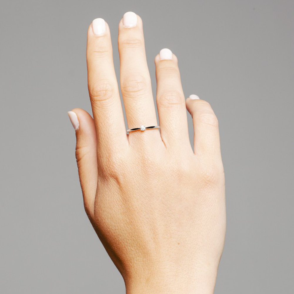 Uma imagem em close-up de uma mão humana contra um fundo cinza. A mão está levantada com o dorso voltado para a frente, mostrando um anel de banda simples usado no dedo anelar. As unhas estão pintadas com um verniz de cor clara