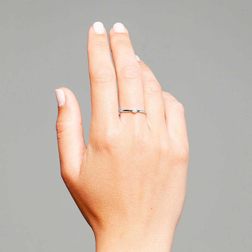 Uma imagem em close-up da mão esquerda de uma pessoa levantada contra um fundo cinza. A mão está posicionada com a palma voltada para dentro e o dorso da mão visível. Os dedos estão ligeiramente afastados e o polegar está encolhido em direção à palma. No dedo anelar, há um anel de banda simples posicionado perto da base do dedo. As unhas estão pintadas com um verniz branco e o tom de pele parece claro.