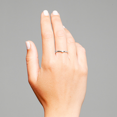 Uma imagem em close-up da mão esquerda de uma pessoa levantada contra um fundo cinza. A mão está posicionada com a palma voltada para dentro e o dorso da mão visível. Os dedos estão ligeiramente afastados e o polegar está encolhido em direção à palma. No dedo anelar, há um anel de banda simples posicionado perto da base do dedo. As unhas estão pintadas com um verniz branco e o tom de pele parece claro.