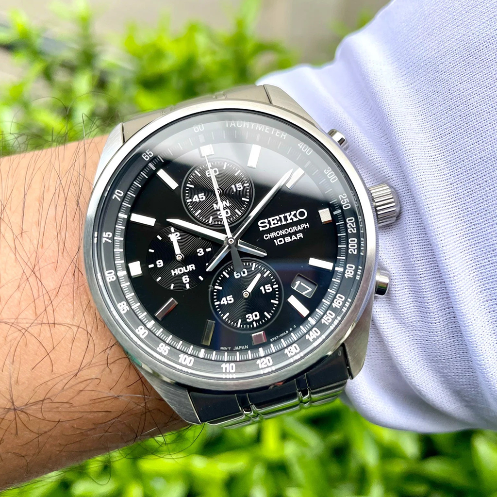 Um relógio cronógrafo Seiko de aço inoxidável é exibido no pulso de uma pessoa. O relógio possui um mostrador preto com marcações brancas, três submostradores e uma janela de data entre as posições das 4 e 5 horas. Os ponteiros são prateados e há uma escala taquimétrica no aro. O fundo mostra folhagem verde desfocada, destacando o relógio como ponto focal.