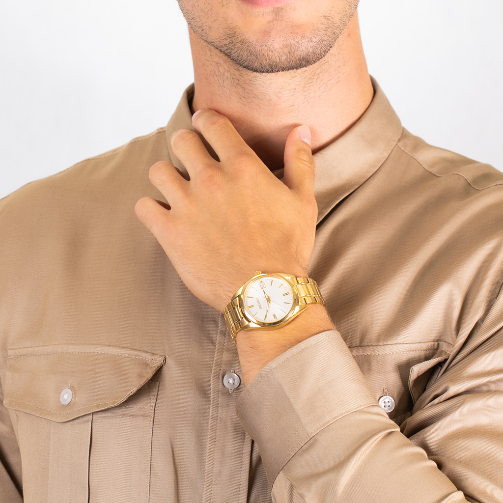 Uma pessoa está usando um relógio de aço inoxidável com tom dourado no pulso esquerdo. O relógio tem um mostrador branco com algarismos romanos pretos indicando as horas e três ponteiros para horas, minutos e segundos. A caixa parece polida e reflete a luz, sugerindo um acabamento metálico. A pessoa está vestindo uma camisa bege com botões visíveis no punho, complementando o estilo elegante do relógio.