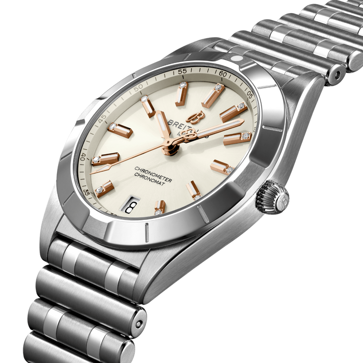 Relógio de pulso Breitling em aço inoxidável, apresentando uma pulseira e caixa prateadas. O mostrador do relógio é branco com marcadores de hora e ponteiros em tom de ouro rosa, e uma janela de data na posição das 3 horas. A marca “BREITLING” está escrita abaixo do marcador das 12 horas, com “CHRONOMETER” e “CERTIFIED” acima do marcador das 6 horas. Há marcadores de minutos ao redor da borda externa do mostrador.