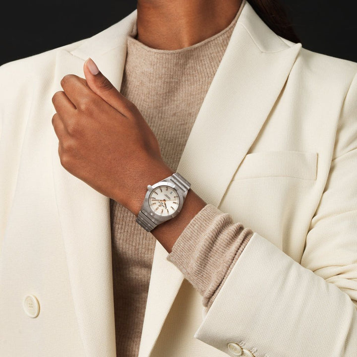 Um relógio de pulso em aço inoxidável com mostrador branco, marcadores de hora pretos e ponteiros no pulso de uma pessoa. A pessoa está vestindo um blazer creme sobre uma blusa de gola alta marrom claro. O foco está no relógio, que parece elegante e sofisticado contra o traje.